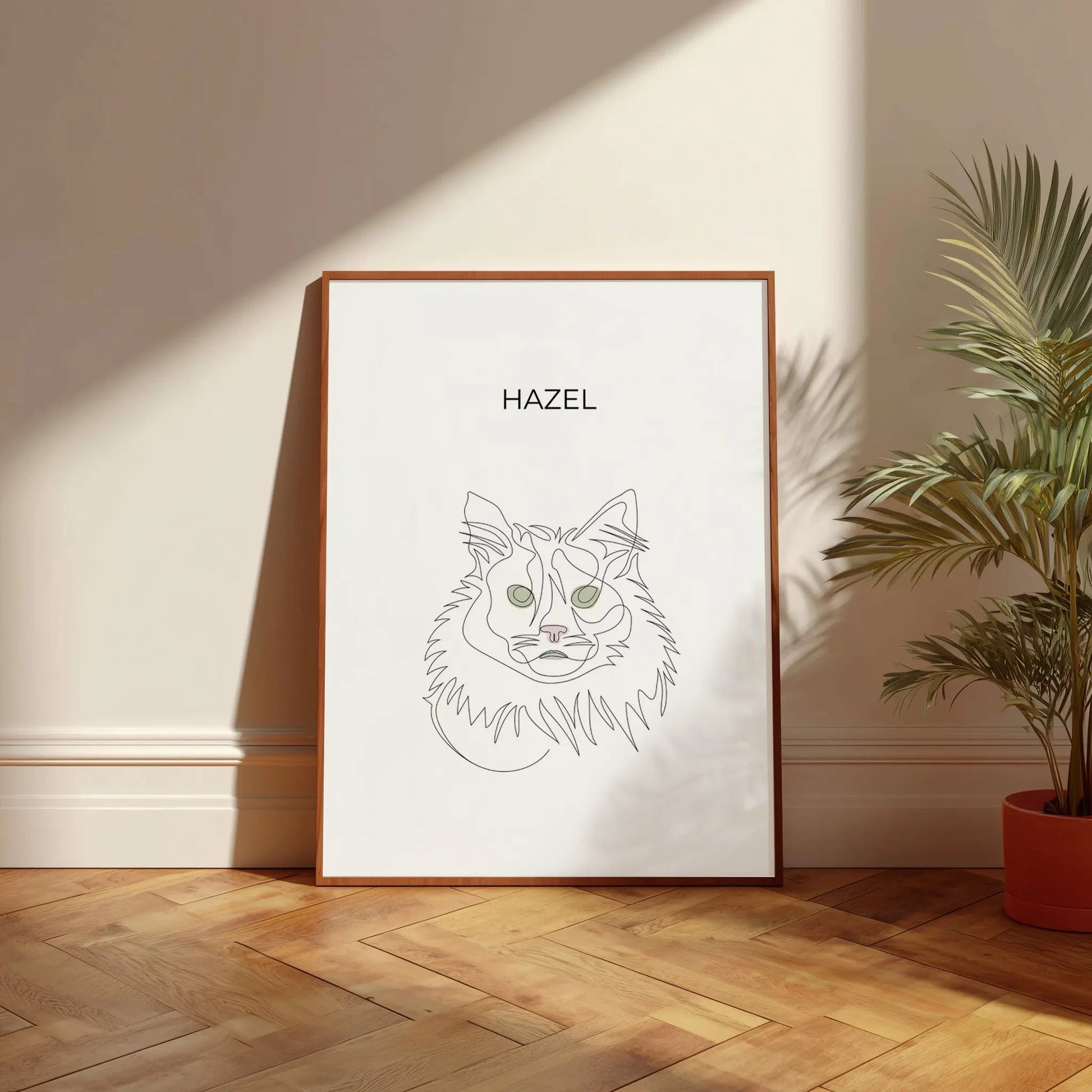 Minimalistisches One Line Art Katzenportrait mit Namen Hazel, individuelles Haustierportrait nach Fotovorlage im modernen Stil als Wandbild mit holzfarbenen Rahmen in 50x70cm vor einer hellen Wand neben einer Pflanze.